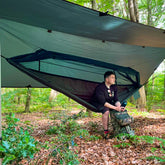 Man relaxing in a DD Frontline hammock under a tarp in a wooded area, showcasing outdoor camping comfort.
